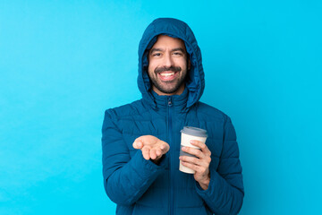 Man wearing winter jacket and holding a takeaway coffee over isolated blue background holding copyspace imaginary on the palm to insert an ad