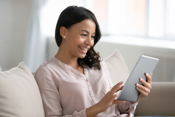 Happy mixed raced woman using tablet for video call at home, resting on couch, looking at screen, smiling. Excited customer shopping online. Student reading electronic book, watching webinar