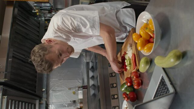 Vertical medium slow-motion footage of male caucasian chef in white jacket cutting fresh organic vegetables on wooden board at restaurant kitchen while watching video recipe via laptop