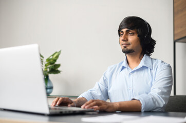 Smiling indian businessman wearing headset is using laptop computer for online communication with customers or colleagues sitting in the modern office