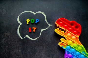 Pop it dinosaur toy rainbow colors on a black background with multicolored letters and the inscription - Pop it in a speech bubble