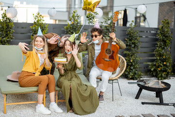 Friends in face masks having a birthday party, making selfie with a cake or having video call on phone while sitting together at backyard outdoors