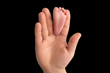 Baby feet in father's hand. The tiny foot of a newborn baby between the fingers of a parent's hand. Open palm and small toes. Black studio background.