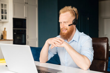 Smiling redhead male freelancer wearing headset and smart casual wear using laptop for connection...