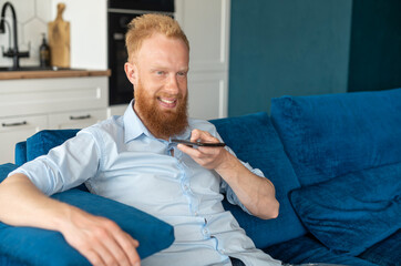 Smiling young redhead man using convert voice into text mobile app, holding smartphone and talking into a mic. Bearded hipster guy recording and sending voice message, writing sms by voice