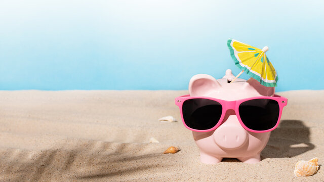 Piggy bank with sunglasses and sunshade umbrella on a sandy beach. Savings for summer vacation or summer sale concept.