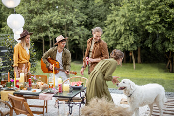 Friends hang out together on a beautiful terrace near the forest, talking and playing with dog and guitar, having great summertime