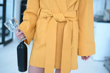 a girl without a face during the day in the kitchen stands in a yellow dressing gown in the model's hand a wine glass and a bottle of wine
