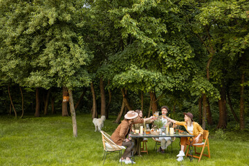 Group of young people having a festive dinner in nature, sitting together by a table on a green...