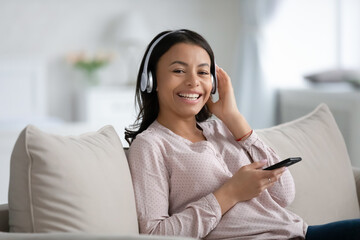 Happy African American woman in wireless headphones listening to music on smartphone, using online app for playing tunes, learning audio lesson, relaxing on couch, looking at camera and smiling