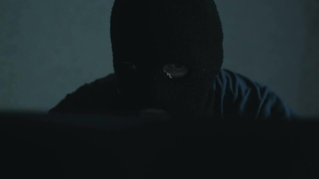 An Internet hacker in a black balaclava sits in a chair and steals data from a laptop. A cyber terrorist wearing a black balaclava hacks into a website and steals important data from Internet users