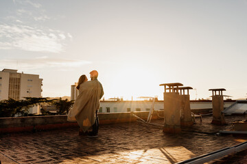 Cute couple standing on rootopg during sunset and hugging each other with blanket
