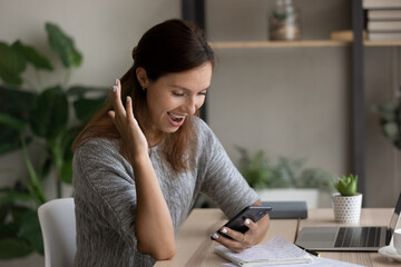 Overjoyed woman looking at phone screen, celebrating success, reading unexpected good news, sitting at work desk, happy young businesswoman received job promotion or reward, business achievement