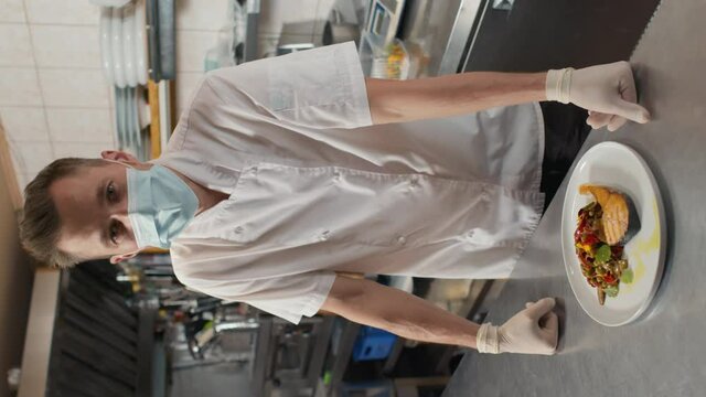 Vertical slowmo medium portrait of experienced male chef in white jacket and protective mask serving plate with prepared dish on table looking at camera