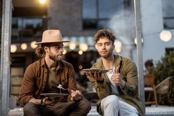 Two male friends eating sweet cakes on a porch of the country house on dusk outdoors. Fun summer time in a country house, male friendship