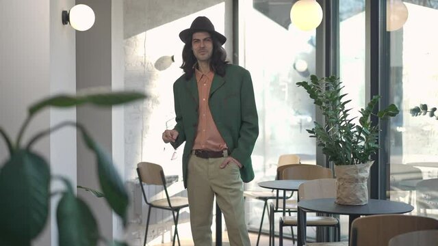 Portrait of stylish 1990s man taking off and putting on sunglasses smiling standing in coffee shop. Confident Caucasian narcissist posing indoors on sunny spring summer day. 1980s lifestyle