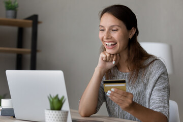 Overjoyed woman holding credit card, looking at laptop screen, excited by success, great sales or money refund, happy satisfied young female customer making secure internet payment, checking balance