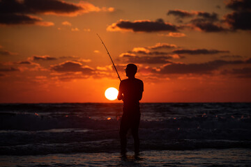 A fisherman standing in the water, holding his rod, at sunset with cloudy sky.