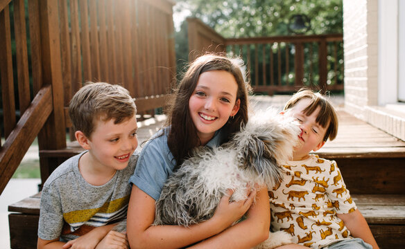 Siblings Sitting Together Outside With Dog, Who is Licking the Youngest Boy's Face