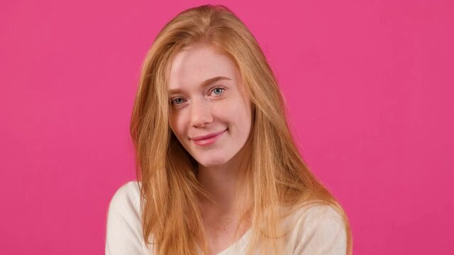 Pretty young red haired woman with freckles looking at camera with a smile and flirting, isolated on pink background.