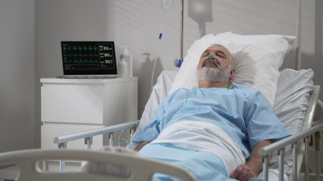 An elderly patient wakes up coming out of a coma. Open your eyes while lying on a bed in a hospital connected In the Hospital Sick Male Patient Sleeps on the Bed, .
