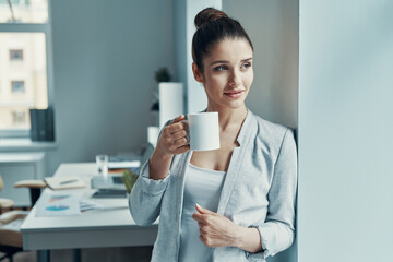 Beautiful young woman in smart casual wear looking away a and holding cup