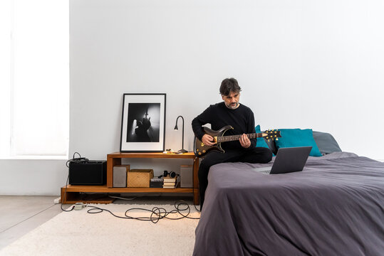 Focused man with laptop playing guitar in bedroom