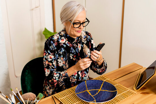 Portrait of senior woman using her smartphone and repairing a broken plate at home