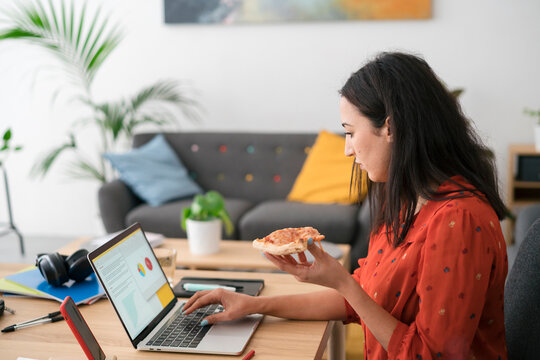 Serious young Arab woman working distantly on laptop and eating pizza at home