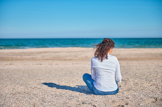Rear view of a young woman in white shirt and blue jeans sitting on the sand in the beach on a beautiful sunny day