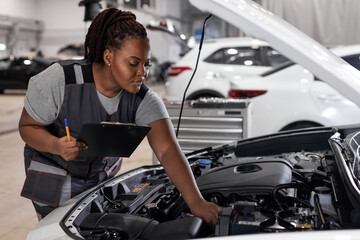 Fototapeta na wymiar car service, job and profession concept. young african female worker in gray uniform with clipboard and pen writing over auto repair shop on background
