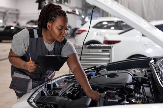 car service, job and profession concept. young african female worker in gray uniform with clipboard and pen writing over auto repair shop on background