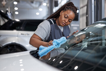 Serious Female afro auto mechanic cleaner is cleaning car window glass with blue rag, side view on...
