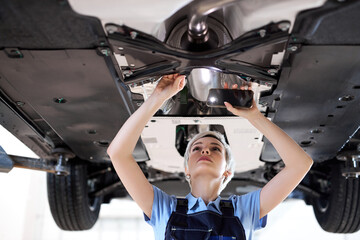 The concept of feminism and women's equality. woman in overalls in auto service. short haired young caucasian woman shines a lantern at the bottom of the lifted car, concentrated and focused