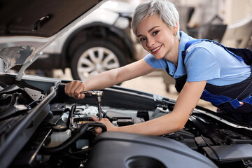 Woman under the hood of car. woman in uniform mechanic repairing a car in auto service. portrait of young short haired hardworking female looking at camera, enjoying work with automobile, vehicle.
