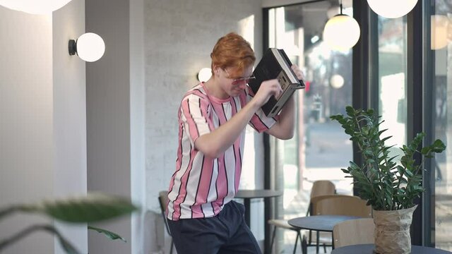 Cheerful redhead Caucasian young man dancing in slow motion to music from vintage tape recorder. Portrait of positive retro guy having fun in coffee shop indoors. 1980s lifestyle and hobby