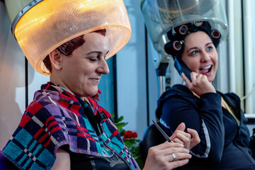 Two female friends at the hairdresser in a beauty salon sitting under a vintage hood hair dryer, talking, gossiping, and laughing while doing their nails. Retro style and fashion. Selective focus.