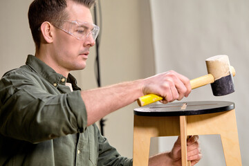 young caucasian confident woodworker making handmade wooden chair in woodworking workshop, side view on male in goggles holding hammer, using instruments tools for carpentry
