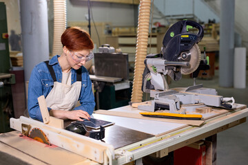 Side view on confident female joiner using digital tablet while working in carpentry workshop, redhead woman in apron and goggles is concentrated, thinking