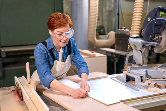 Redhead female carpenter working as wood designer in small carpentry workshop. Young business woman handcrafting piece of timber and designing new house furniture. Entrepreneurs concept lifestyle
