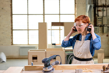 Woman carpenter wearing headset before woodworking, cutting wood using power saw in workshop. Redhead Young female carpenter in safety equipment making furniture, side view