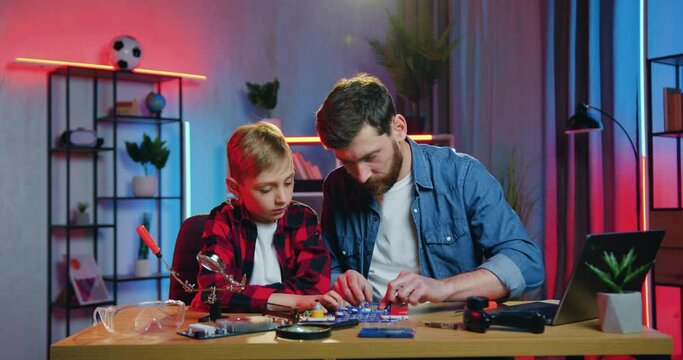 Handsome caring confident positive bearded father helping to assemble the small parts of constructor together with his nice interested teen son at home