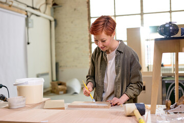 Nice Caucasian Carpenter Handywoman Painting Wooden Plank In Workshop, Portrait Of Redhead Female...