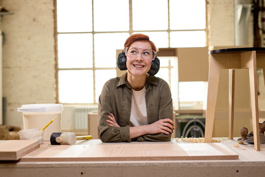 Portrait of positive female carpenter wearing headset and goggles in workshop, looking at camera having pleasant smile, after hard working day. copy space. woodworking, carpentry concept