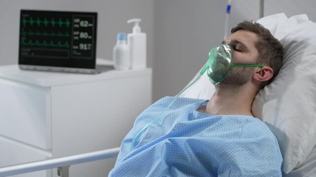 Caucasian man with oxygen mask on lying in bed with white linen, sleeping disturbingly, moving head. Portrait of a man in an oxygen mask who lies on a bed in a hospital.