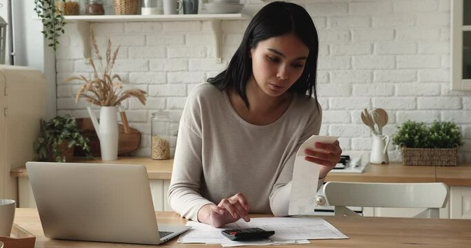 Serious young Asian woman sit at table in kitchen calculate expenses, holds cashier check sales slip use calculator makes calculations, manage personal finances, e-pay household bills services concept