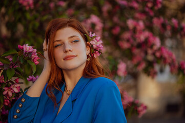 Young elegant redhead freckled woman wearing blue blazer posing near pink tree blossom. Model looking aside. Copy, empty space for text 