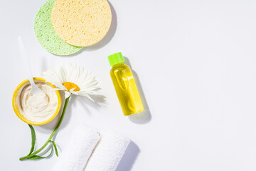 Spa accessories clay face mask, scrub sponge, facial lotion with herbal extract of chamomile flowers, towels, essential oil on white background, top view. Woman care cosmetics with natural ingredients