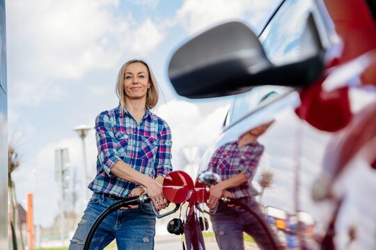 Pretty blonde woman is filling her red car with gasoline at a gas station.