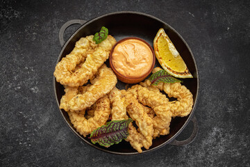 Golden rings of squid fries in a round frying pan with sauce, herbs and lemon on a dark background without decorations
